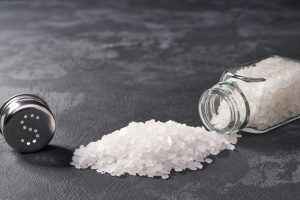 Pile of salt and salt shaker on black stone background. Horizontal composition.